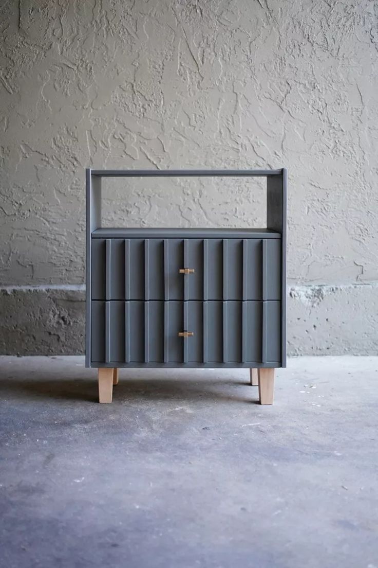 a grey cabinet with three drawers and two wooden legs in front of a concrete wall