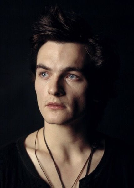 a young man with blue eyes is posing for a photo in the dark, wearing a black shirt and necklace