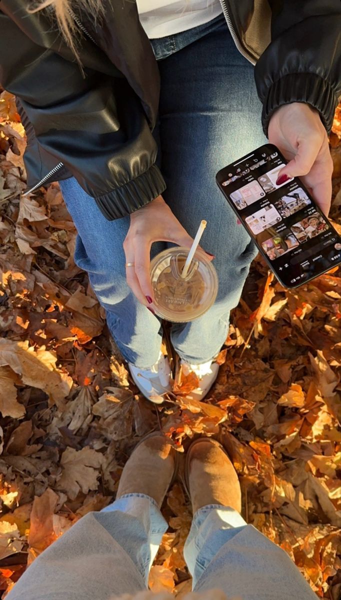 a person sitting in leaves holding a drink and cell phone