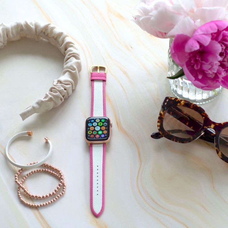 an apple watch, sunglasses, and bracelet on a white table with pink flowers in a vase