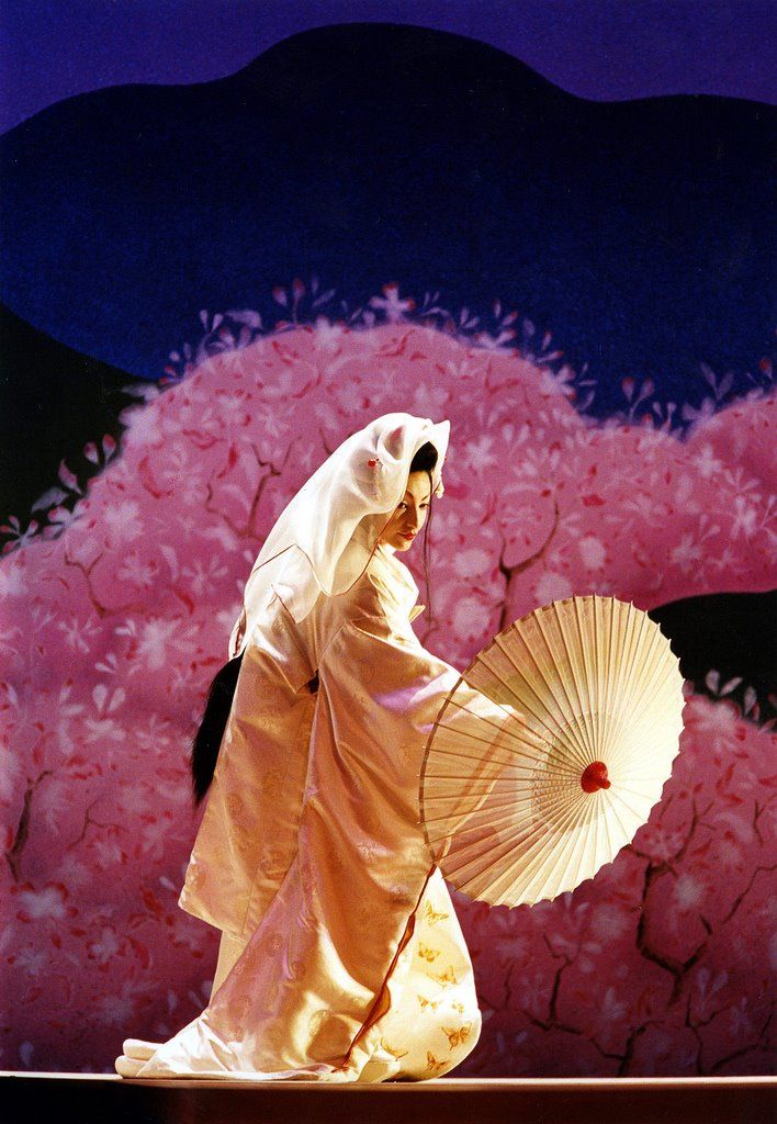 a woman dressed in traditional japanese garb and holding an umbrella walks past pink flowers