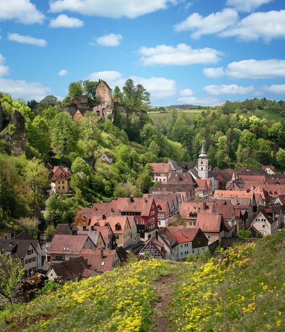 the village is surrounded by lush green trees and yellow wildflowers, with a church in the background