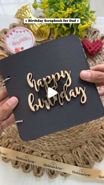 two hands holding a black birthday card with the words happy birthday written on it in wooden letters