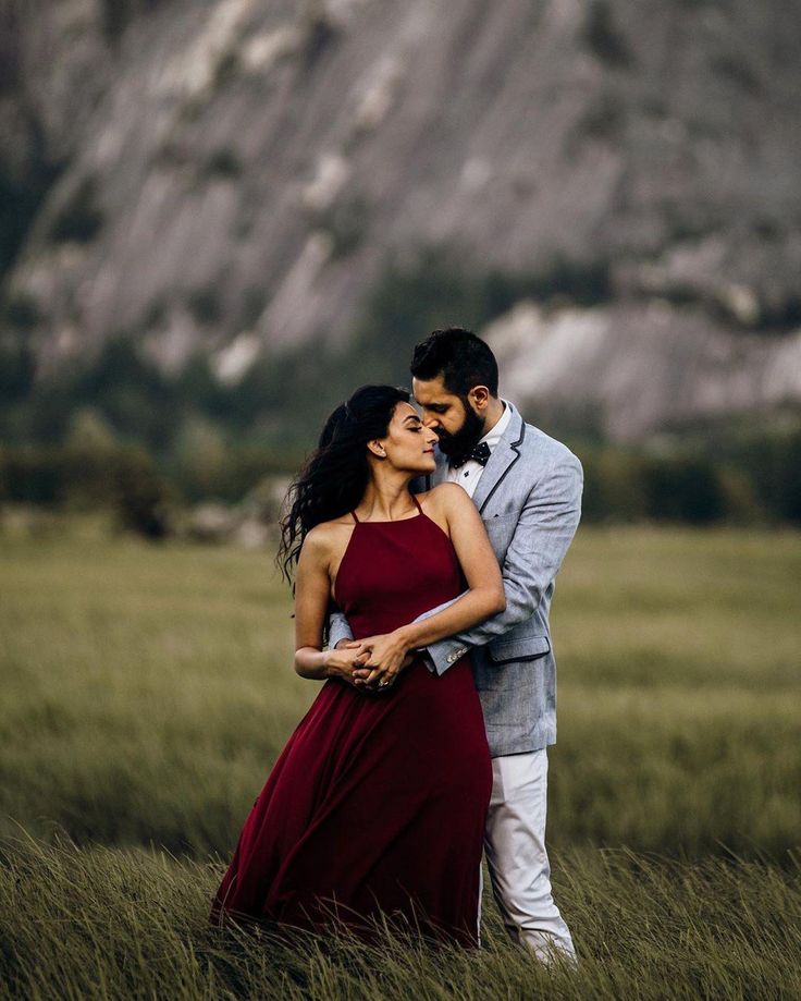 a man and woman standing in tall grass with mountains in the background