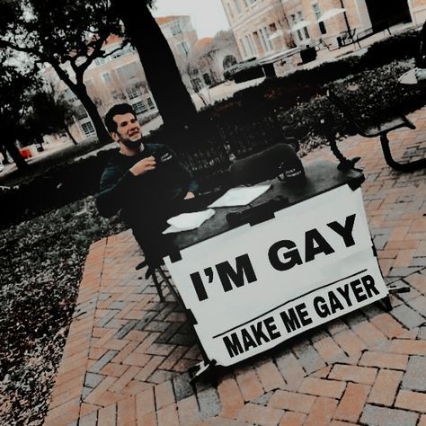 a man sitting on a bench with a sign that says i'm gay make me gay