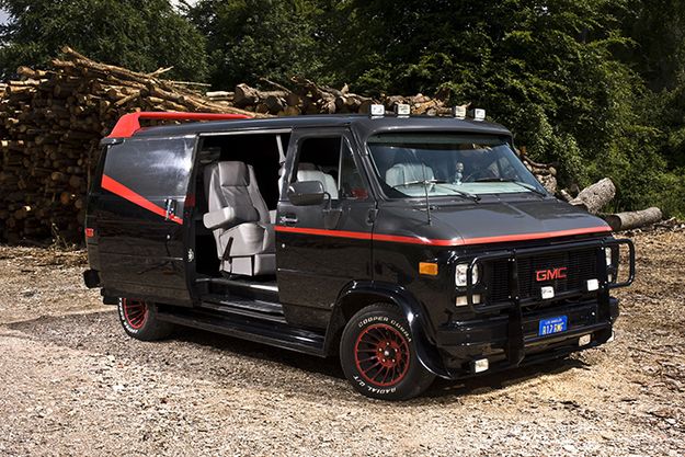 a black van with red trim parked in front of a pile of logs and trees