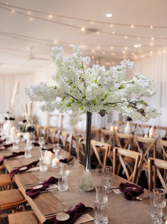 an arrangement of white flowers in a tall vase on top of a long table set with wine glasses