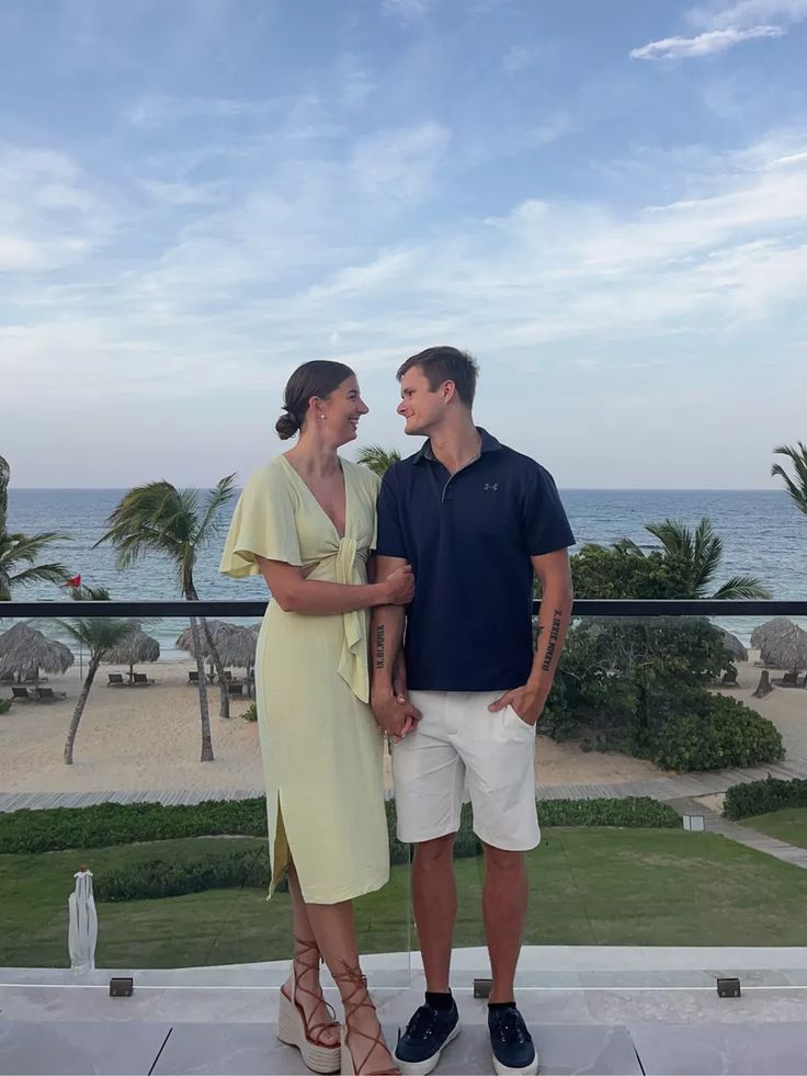 a man and woman standing next to each other on a balcony near the ocean with palm trees