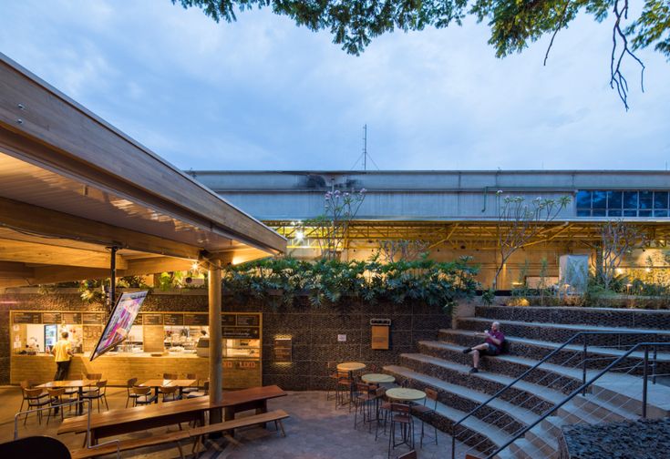 an outdoor seating area with tables and benches at dusk, next to a building that has stairs leading up to the roof