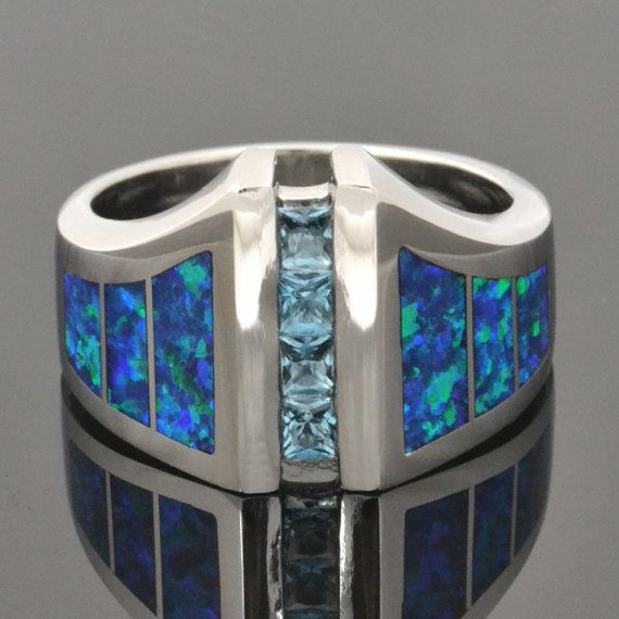 Lab created opal ring with blue topaz accents set in sterling silver. This man's ring is inlaid with bright blue-green synthetic opal that is is a great match with sparkling blue topaz. There are 3 pieces of lab created opal inlaid on each side of the ring. The four princess cut blue topaz total .60 carats and are channel set down the center of the ring. #labopalring Four Princess, Survival Hacks, Rings Opal, Natural Opal Ring, Inlay Jewelry, Mens Ring Sizes, Synthetic Opal, Engagement Rings Opal, Large Ring