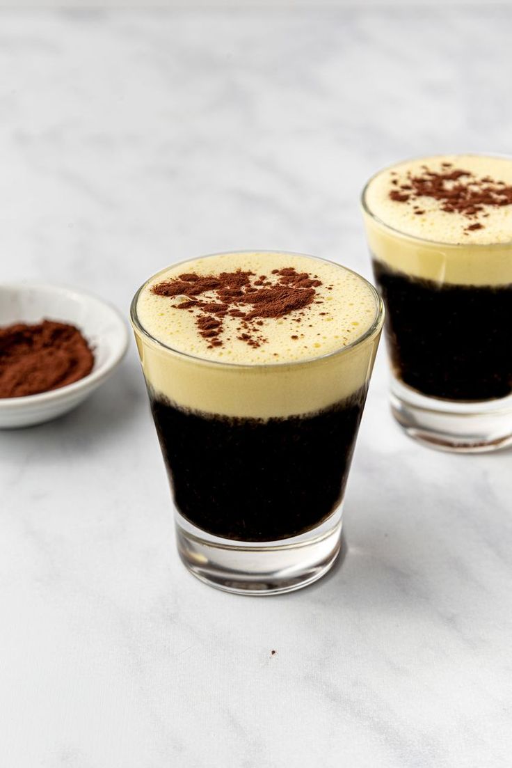 two shot glasses filled with liquid and topped with chocolate powder, on a marble surface