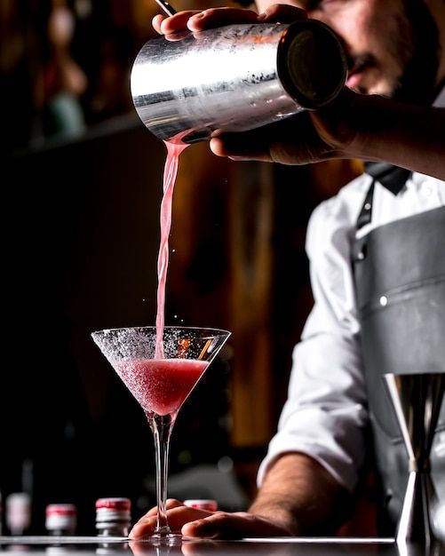 a bartender pouring a drink into a martini glass at a bar with red liquid coming from the top