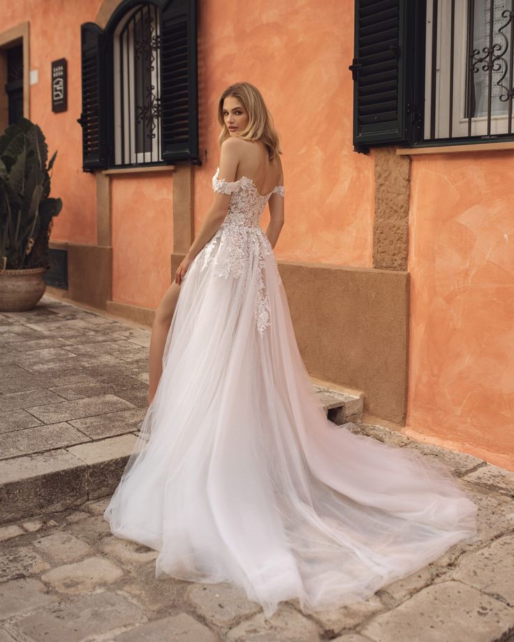 a woman in a white wedding dress standing on the steps near an orange building with black shutters