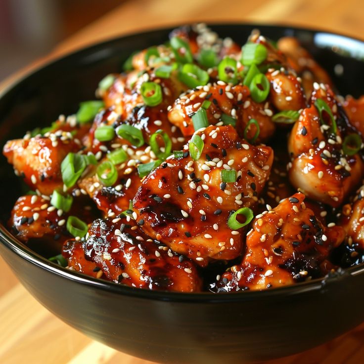 a black bowl filled with chicken covered in sesame seeds and garnished with green onions