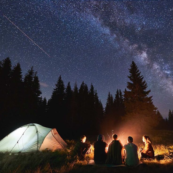 four people sitting around a campfire under the night sky with their tent in the foreground