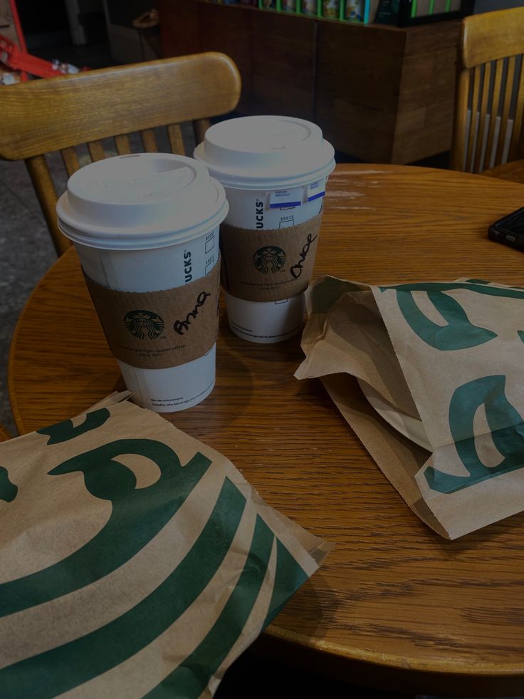 two cups of coffee sitting on top of a table next to some paper bags and a laptop