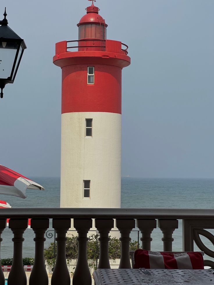 a red and white light house sitting next to the ocean with an umbrella over it