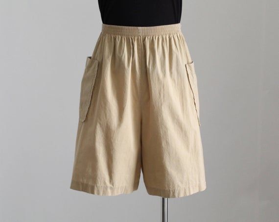 Vintage 80's high waist cargo shorts in beige. Elastic waistband. Long length. Loose fit. Unlined. Lightweight material.Waistband unstretched* - 26.5" --- 67.3 cmHips** - 49.5" --- 125.8 cmInseam - 7.5" --- 19 cmOutseam - 20" --- 51 cmRise - 14.25" --- 36 cmLeg opening - 30" --- 76 cmNote *Since the waistband is elastic, it best fits someone who's waist is at least a few inches larger than 26.5".**Leave some wiggle room for your hips so you can move around comfortably, at least 3" or 7.5 cm.– Be Vintage Summer Bottoms With Cargo Pockets, Vintage Cargo Bottoms For Summer, Vintage Shorts With Side Pockets For Summer, Beige Pants With Pockets And Short Inseam, Short Length Cargo Pants, Short Cargo Bottoms For Workwear, Vintage Summer Cargo Shorts With Pockets, Vintage Summer Shorts With Cargo Pockets, Vintage Short Bottoms For Workwear