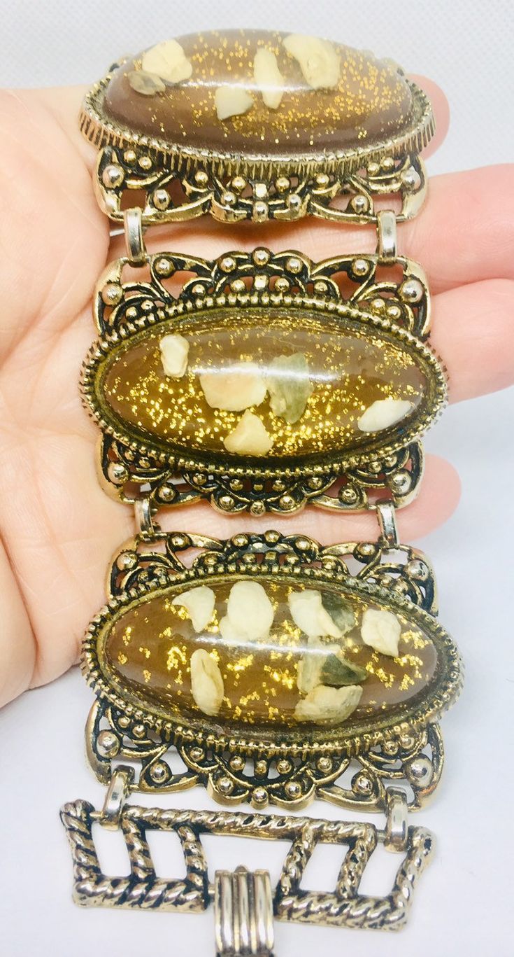 Fabulous vintage extra wide Lucite confetti glitter bracelet with imbedded pebbles. Long lucite cabochons set in ornate antiqued gold tone metal settings. Brown/copper and gold glitter color perfect for fall. Bracelet is in excellent vintage condition with minimal age appropriate wear... if any. It measures 7 1/2 inches long by 2 inches wide. Clasp secure.   Thanks for looking. Antique Gold Cabochon Bracelets, Antique Gold Bracelets With Cabochon, Collectible Gold Bracelets With Cabochon, Gold Metal Bracelets With Cabochon, Gold Metal Bracelet With Cabochon, Bohemian Gold Oval Bracelets, Gold Oval Bohemian Bracelets, Antique Jeweled Gold Bracelets, Vintage Resin Jewelry For Party