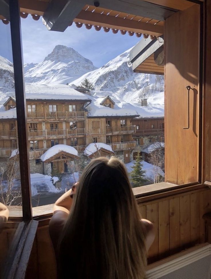 a woman sitting in front of a window looking out at snow covered mountains and buildings