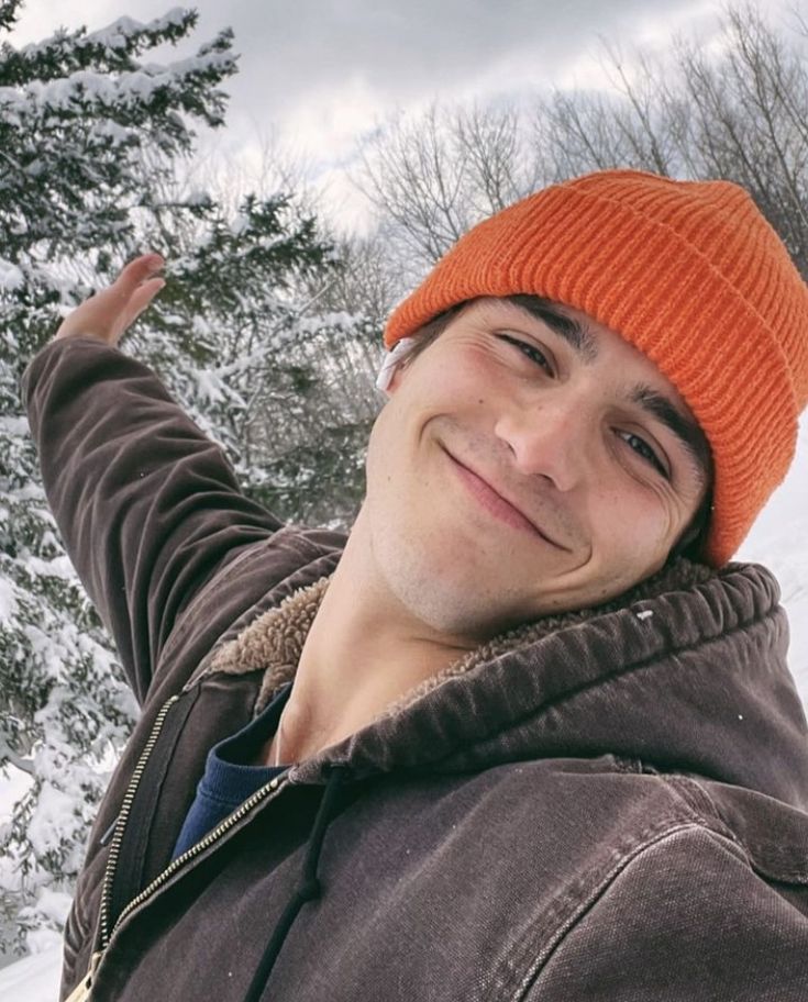 a young man wearing an orange beanie smiles at the camera while standing in front of snow covered trees