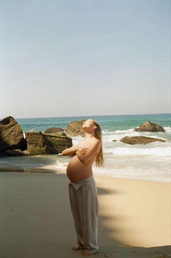 a pregnant woman standing on the beach with her belly exposed, looking up at the sky