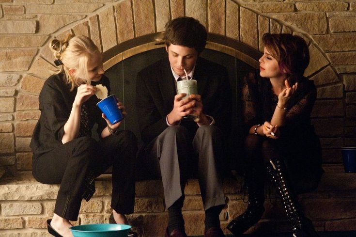 two women and a man sitting on a bench drinking from cups in front of a fireplace