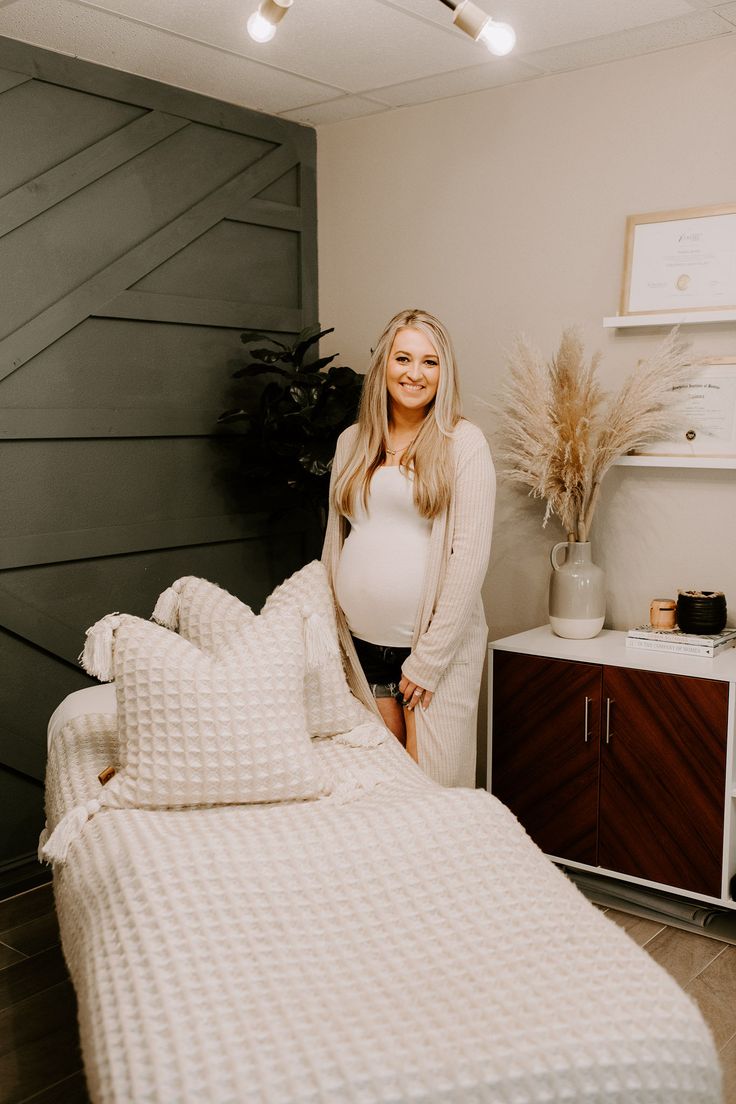 a pregnant woman standing next to a bed in a room with white sheets and pillows