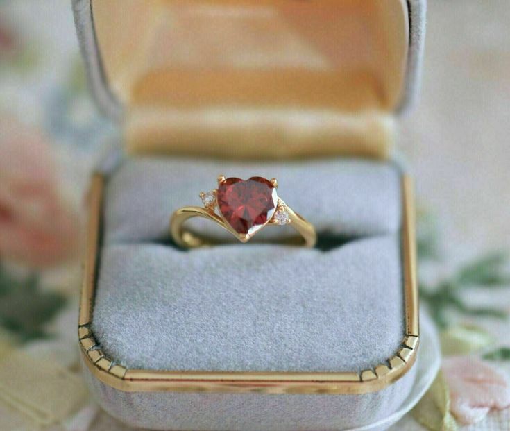 an engagement ring with a heart shaped red stone sits in a box on a floral tablecloth