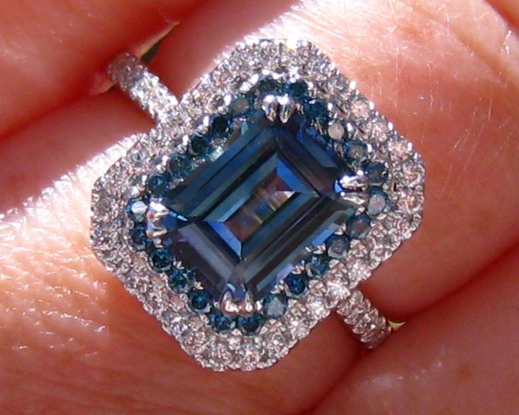 A beautiful and unique engagement ring featuring a teal blue sapphire and blue diamonds in white gold diamond double halo. The center stone is an emerald-cut sapphire, that weighs 1.98 carat and measures 8.5x6.3mm. The sapphire is basic heat only and comes from Sri Lanka. It has a beautiful teal blue color and clean crystal that matches great with blue diamonds. Be sure to check out the sparkle in the video! The sapphire is set in a 14K white gold diamond double-halo ring. The inside halo is don Blue Emerald-cut Halo Ring With Center Stone, Blue Emerald Cut Halo Ring, Blue Sapphire Ring With Radiant Cut And Accent Stones, Gia Certified Blue Sapphire Ring In Platinum, Blue Gia Certified Diamond Halo Ring, Gia Certified Blue Diamond Halo Ring, Blue Radiant Cut Diamond Ring With Accent Stones, Blue Sapphire Ring In Radiant Cut Platinum, Blue Sapphire Ring Radiant Cut For Wedding