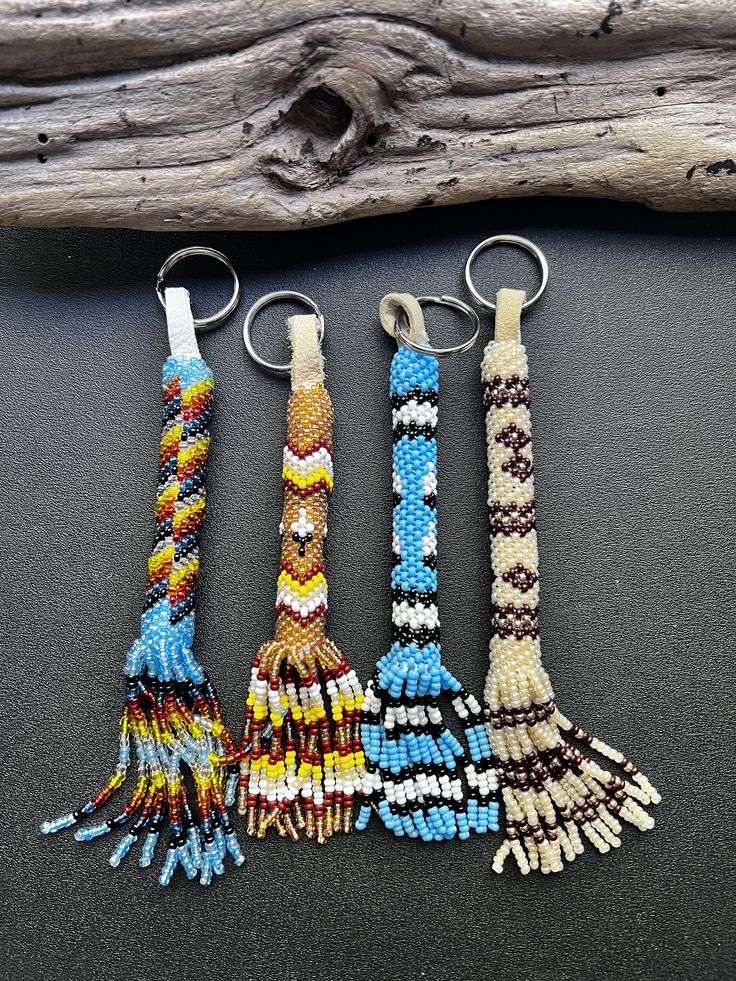 three key chains with beads and tassels hanging from them on a table next to a piece of driftwood