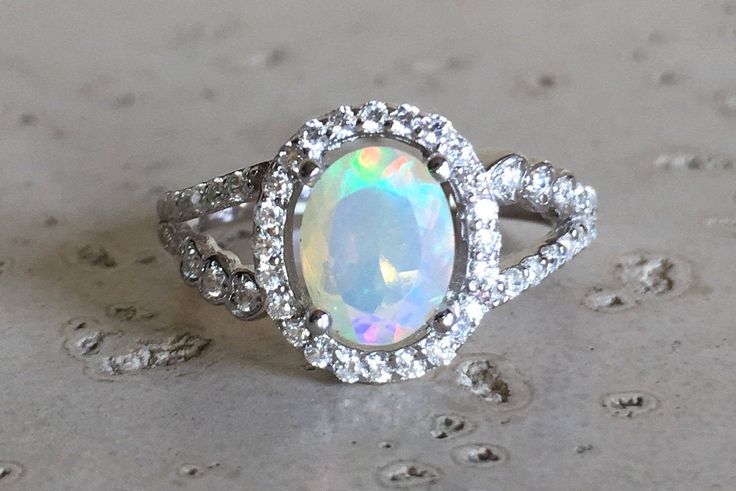 "A lovely Halo Setting featuring an oval-shape genuine top-notch Welo Opal with a magnificent play of color, complemented with a split shank embellished with pave set cubic zirconia handcrafted in Sterling Silver. Wrapped in a box ready for gift-giving.(r-egt-70 ) FINAL SALE Ring Info ----------------- *Opal measure 6mm x 8mm *Opal with Halo measure 11.5mm x 10mm *Ring Shank Width 2mm *Stone height 8mm *Model ring size 5 HOW TO CARE FOR YOUR OPAL --------------------------------------------- Opa Moissanite Opal Engagement Ring, Oval Opal Ring With Gemstone Accents For Anniversary, Oval Opal Ring With Accent Stones, Dazzling Oval Opal Ring, Oval Opal Ring With Halo Setting, Elegant Oval Opal Ring With Gemstone Accents, Dazzling Oval Opal Ring For Anniversary, Oval Promise Ring, Juelz Santana