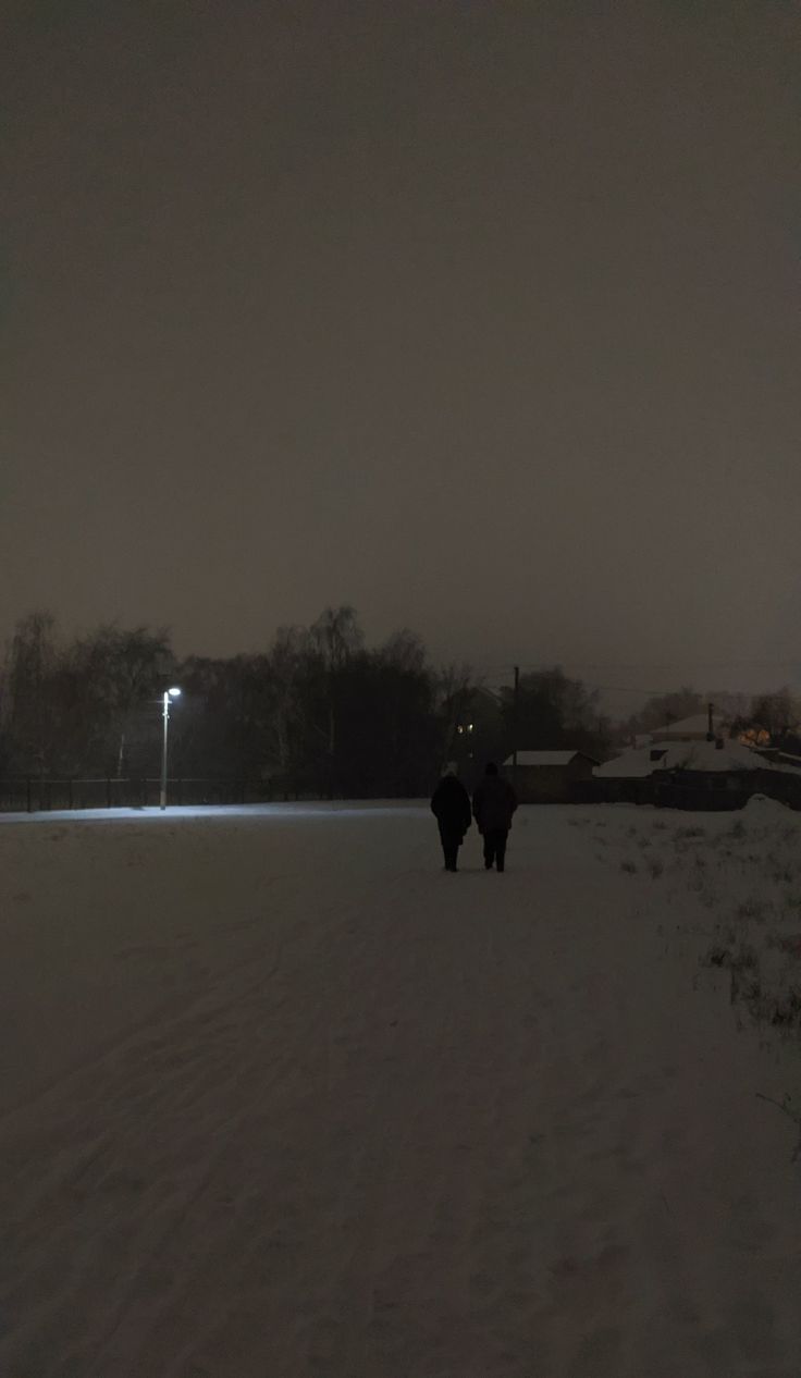 two people walking in the snow at night with lights shining behind them and buildings on either side