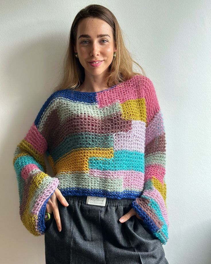 a woman standing in front of a wall with her hands on her hips wearing a multicolored sweater