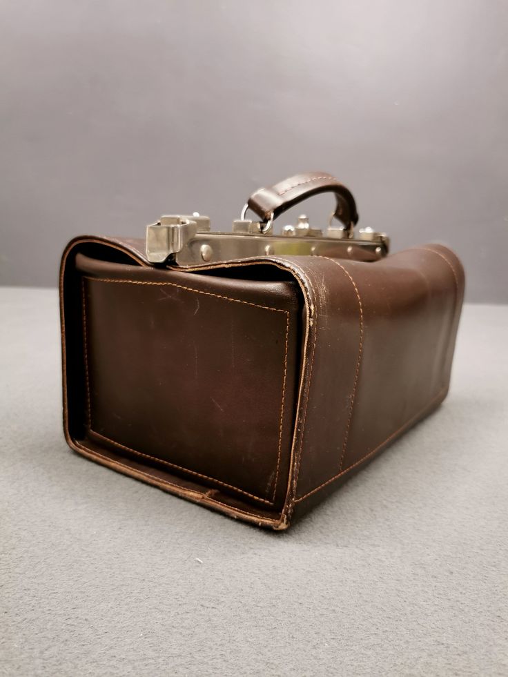 The brown leather bag from the 1960s exudes vintage charm and functionality. This elegant bag likely served doctors or veterinarians of that era to carry their precious tools. With its triangular profile and sturdy leather handles, the bag appeared serious and authoritative. The bag's interior was probably lined with soft material to protect medical or veterinary instruments. This well-designed bag was not only practical but also reflected the professional reputation of its owner. Over time, this bag has become a valuable collector's item. Its quality craftsmanship and authentic vintage appearance make it special. Whether used as a functional piece or as a decorative element in an interior, the brown leather bag from the 1960s leaves a strong impression and carries with it a story of its m Retro Leather Box Bag For Travel, Retro Leather Box Bag With Top Handle, Retro Leather Top Handle Box Bag, Vintage Office Bags With Brass Hardware, Vintage Leather Rectangular Briefcase, Vintage Cognac Briefcase For Everyday, Vintage Cognac Briefcase, Retro Leather Satchel Box Bag, Vintage Leather Satchel With Top Handle