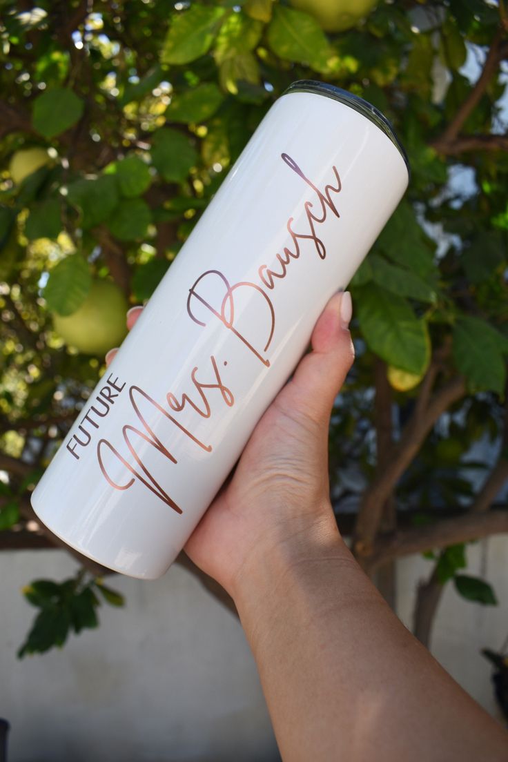 a hand holding a white coffee cup with writing on the bottom and inside, in front of a tree