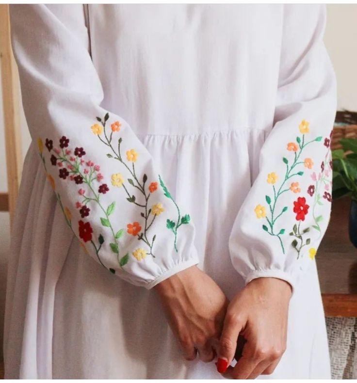 a woman in a white dress is holding her hands together with flowers embroidered on the sleeves