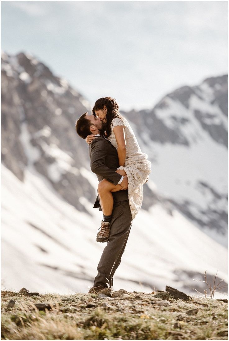 a man holding a woman in his arms on top of a snow covered mountain with mountains in the background