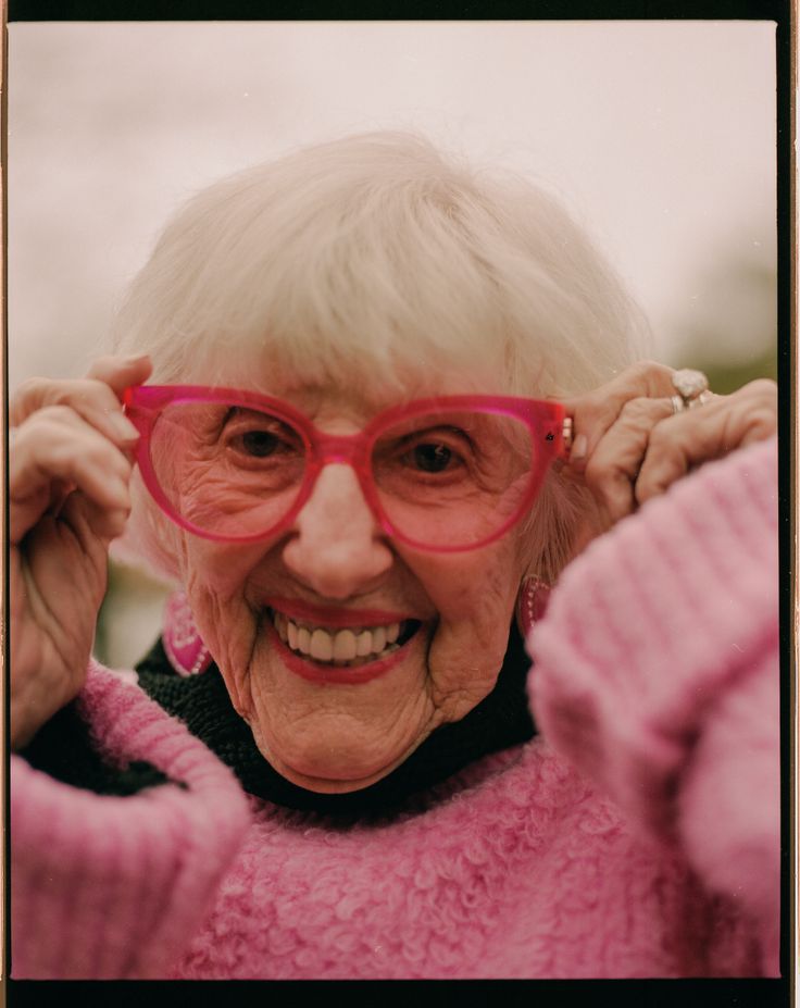 an older woman wearing pink glasses smiling at the camera with her hand on her head