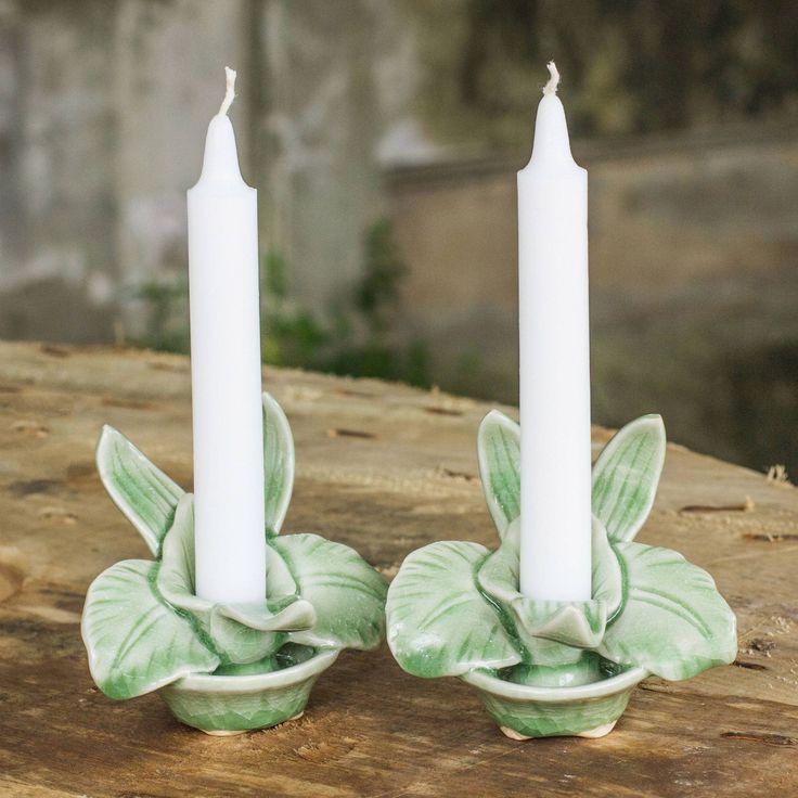 two white candles sitting on top of each other in front of a wooden table with green leaves