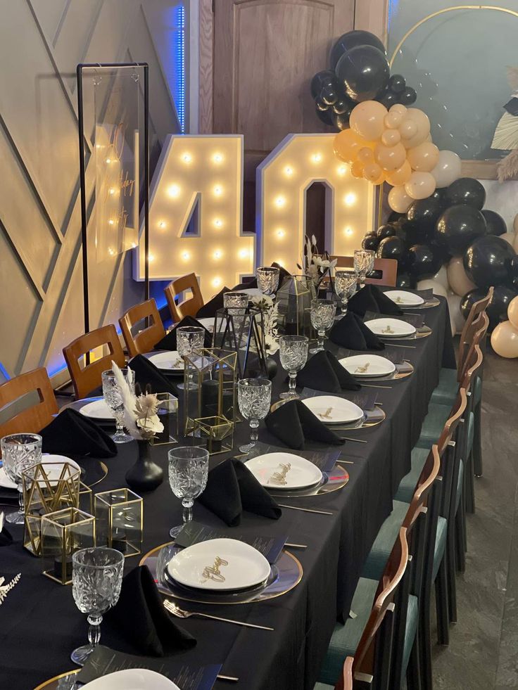 the table is set with black linens and white plates, gold rimmed glassware, and balloons