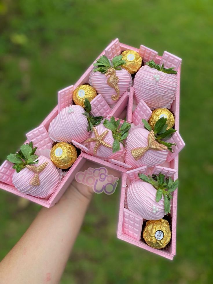 a hand holding a pink box filled with chocolate covered strawberries and gold foil wrapped strawberries