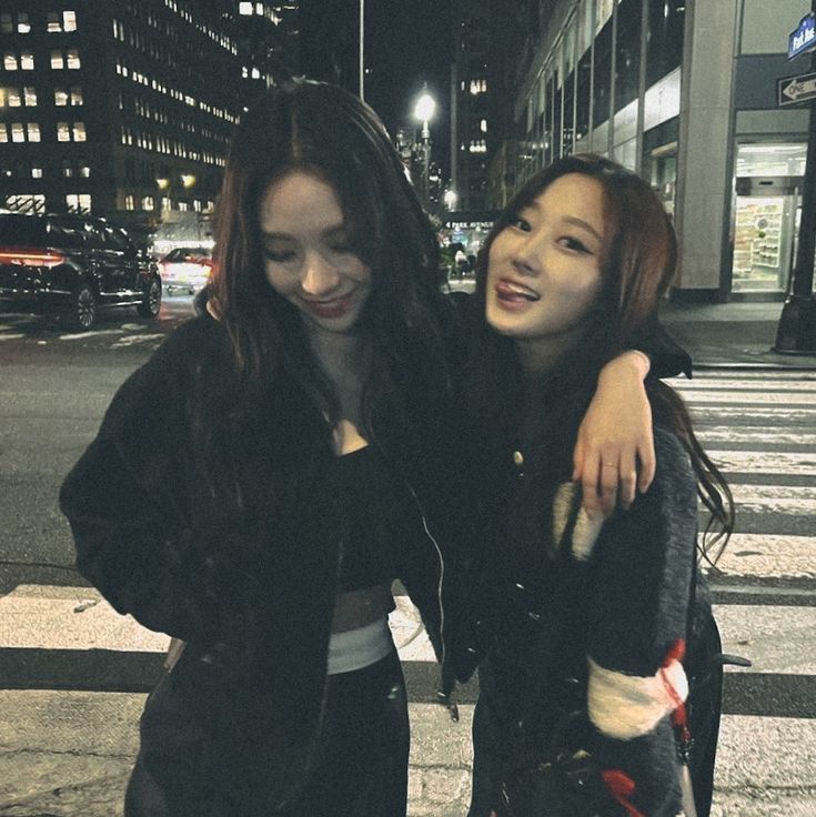 two young women standing next to each other on a city street at night with buildings in the background