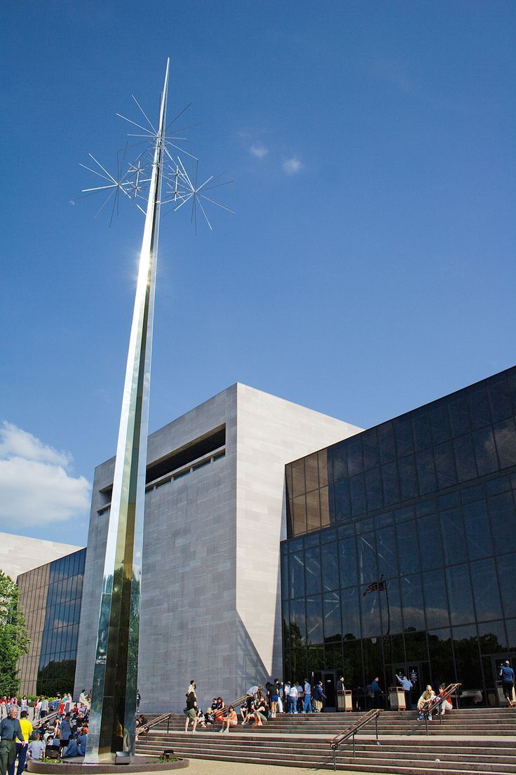 a tall metal pole in front of a building with people sitting on the steps outside