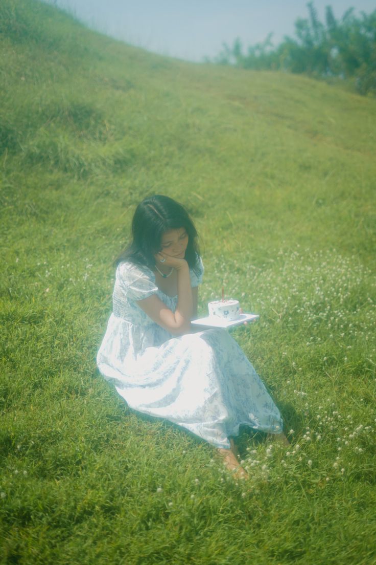 a woman in a white dress sitting on the grass with a tea cup and saucer