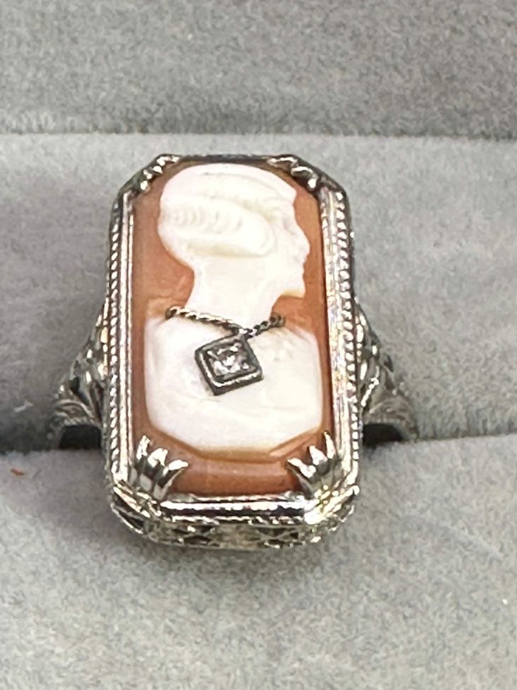 For your consideration, a 14K Art Deco, white gold cameo ring. This beautiful ring is hallmarked as 14K and guaranteed to be such. There is no makers mark. The cameo is a carved shell that is prong set. The setting measures 20.8 mm x 11.6 mm. The cameo's subject is wearing a traditional 1920s wavy hairstyle and is wearing a white gold chain with a 2-point diamond pendant.The setting is very typical of art deco styling with a floral motif all the way around the top of the ring  that continues around the sides of the ring. This fabulous ring is a size 7 1/2 weighs 3.27 g. It is a fabulous piece of history with a lot more history to be made! Fine Carved Diamond Jewelry, Carved Diamond Fine Jewelry, Cameo 14k Gold Ring, Art Deco White Gold Jewelry With Cabochon, 14k Gold Cameo Ring, Art Deco Cameo Ring For Collectors, Antique Cameo Ring Jewelry, Art Deco Cameo Ring Collectible, Art Deco Cameo Collectible Rings