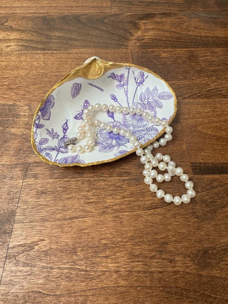 a white and purple flowered dish with pearls hanging from it's side on a wooden floor