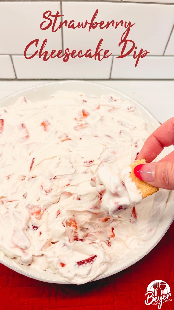 strawberry cheesecake dip on a white plate with a hand holding a cracker over it