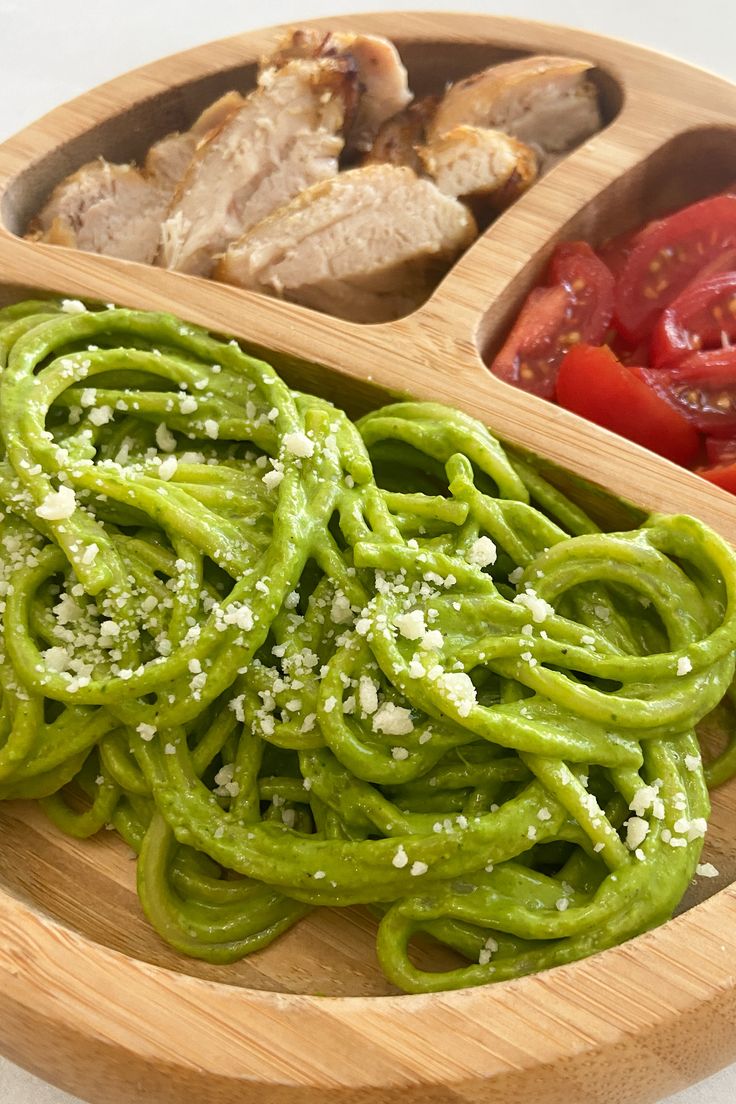 a wooden bowl filled with green pasta and tomatoes next to meat on a cutting board