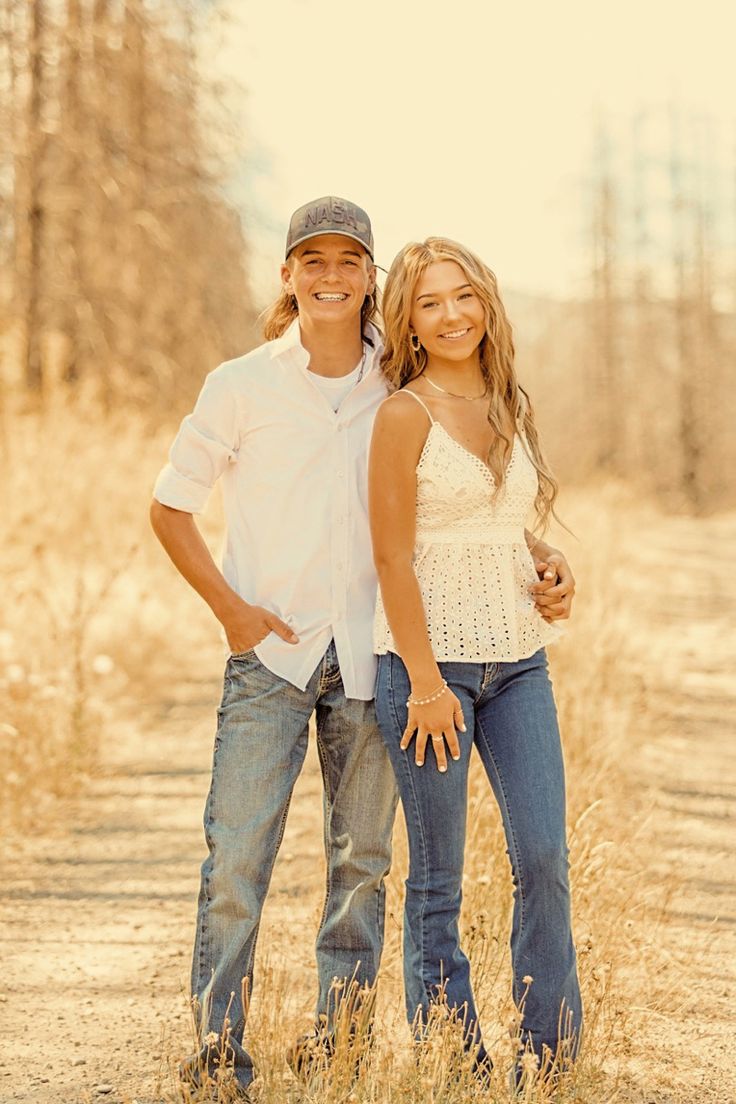 a man and woman standing in the middle of a dirt road with trees behind them